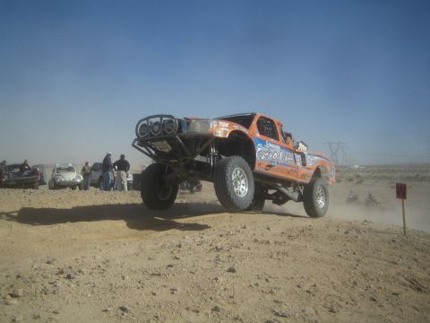 1990 Ford Bronco - Action photo of an orange off-road race truck catching air over a desert berm with spectators watching; not a technical document.