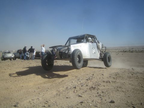 1990 Ford Bronco - Photograph of a silver tube-frame off-road race car driving over a desert berm with onlookers nearby.