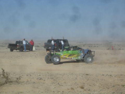 1990 Ford Bronco - Photograph of a green tube-chassis off-road race buggy racing past spectators and pickup trucks in the desert.