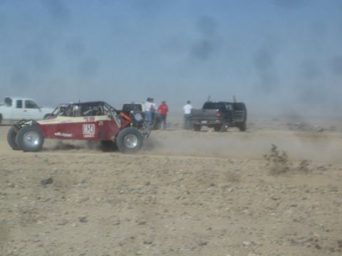 1990 Ford Bronco - Photograph of a red tube-frame off-road race buggy racing through dust past parked trucks.