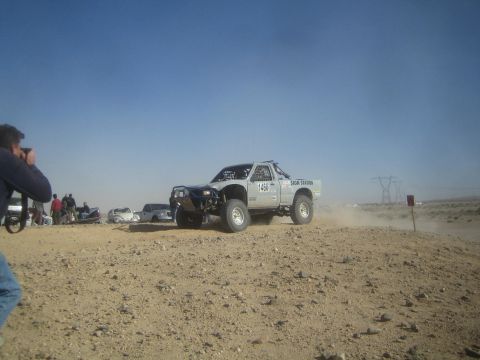 1990 Ford Bronco - Photograph of a white off-road race truck catching air on a rocky desert race course as a photographer shoots.