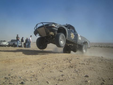 1990 Ford Bronco - Photograph of a dark off-road race truck launching off a berm with front wheels in the air.
