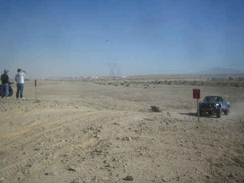 1990 Ford Bronco - Photograph of an off-road race vehicle on a wide desert course with spectators and a course marker.