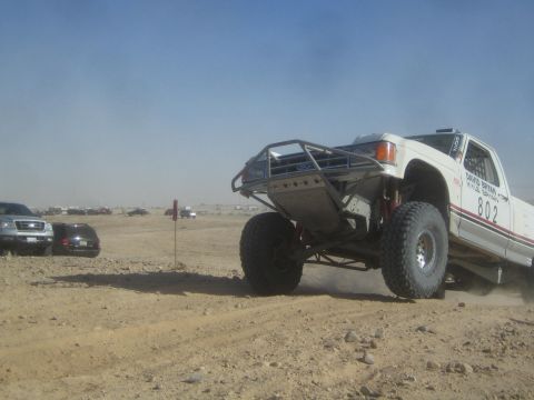 1990 Ford Bronco - white off-road race truck numbered 802 climbs over a desert berm with its front end raised, parked cars visible in the background.