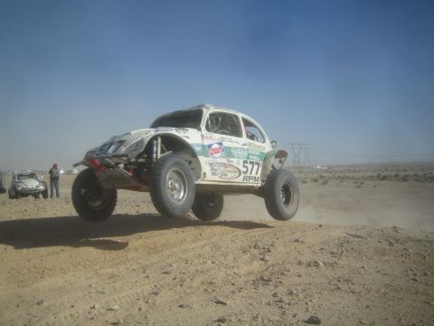 1990 Ford Bronco - white Baja Bug VW Beetle racer numbered 577 launches over a desert rise, kicking up dust with spectators in the background.