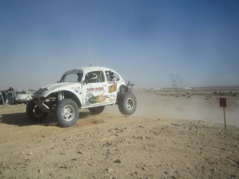 1990 Ford Bronco - white Baja Bug VW Beetle racer with flame graphics and number 557 speeds across a dusty desert course as spectators watch from the left.