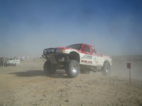 1990 Ford Bronco - red and white off-road race truck numbered 1472 runs across a dusty desert course beside a red W course marker.