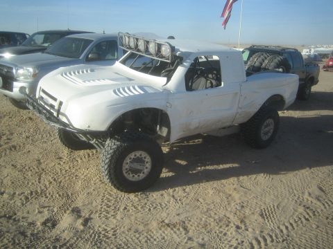 1990 Ford Bronco - white fiberglass-bodied prerunner truck with a roof-mounted light bar sits on desert sand near pickups and a flag at a staging area.