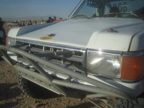 1990 Ford Bronco - close-up of a white off-road truck's front end showing a tubular bumper, chrome grille, and headlights at a desert staging area.