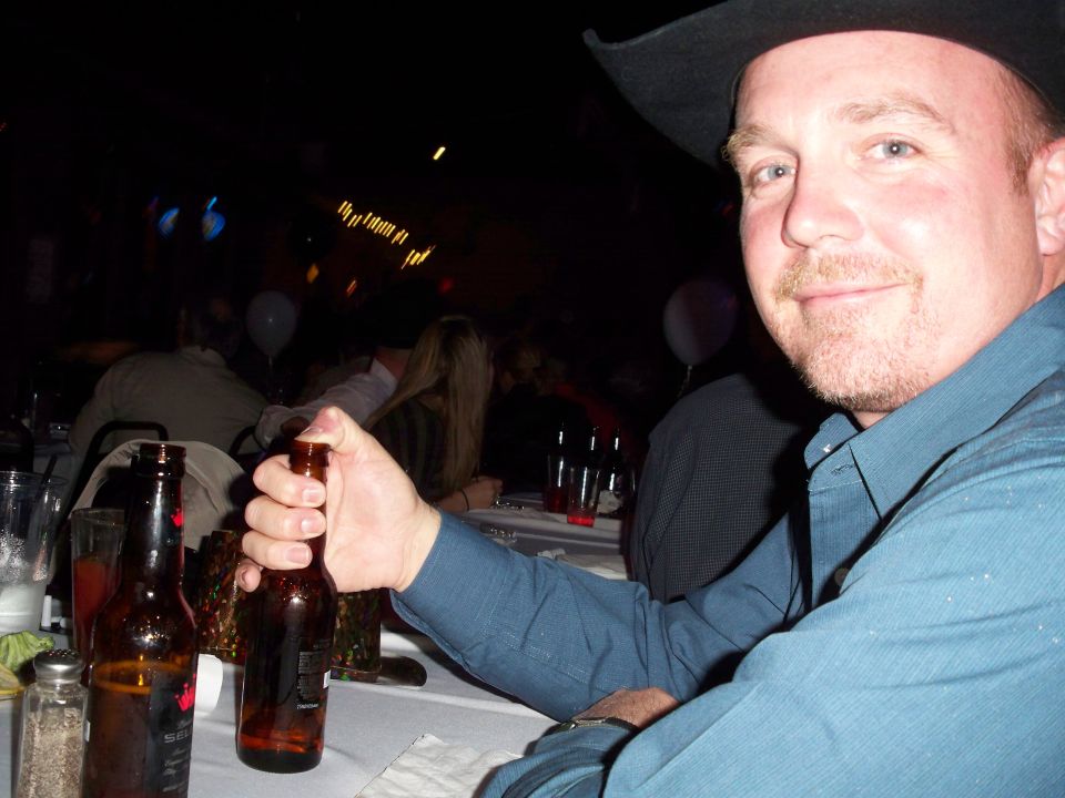 Man in cowboy hat at table, holding a beer bottle. Two beer bottles on table