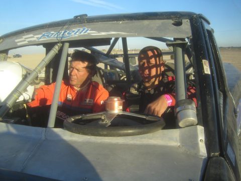 1990 Ford Bronco - driver in an orange suit and a passenger sit in the open tube-frame cab of an off-road race truck out in the desert at sunset.