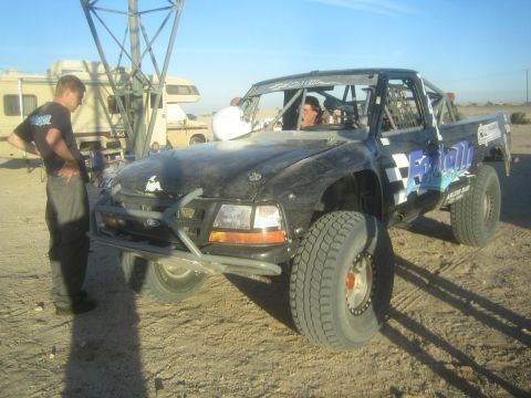 1990 Ford Bronco - black off-road prerunner truck with checkered graphics sits in a desert pit area near an RV as a man stands beside it at golden hour.