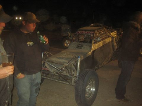 1990 Ford Bronco - Two men holding drinks stand beside a long tube-frame off-road buggy parked in a dark lot at night.
