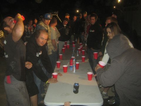 1990 Ford Bronco - crowd gathers around a folding table lined with red cups and beer cans playing a drinking game at night.
