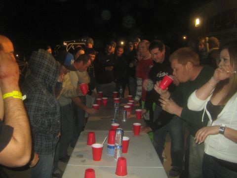 1990 Ford Bronco - Men and women lean over a folding table covered in red cups, competing in a flip-cup style drinking game at night.