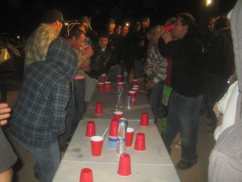 1990 Ford Bronco - crowd surrounds a long folding table stacked with red cups and beer cans during a nighttime drinking game.