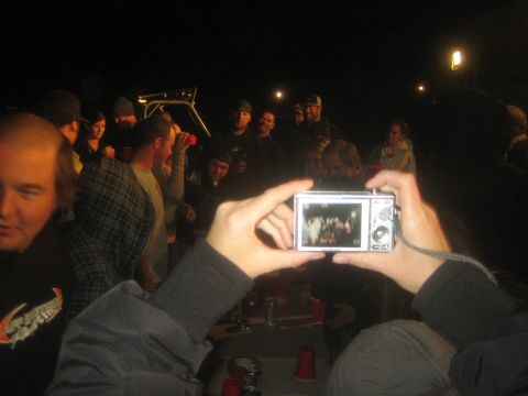1990 Ford Bronco - person holds up a small silver digital camera to photograph a crowd gathered at a dark outdoor party.