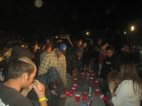 1990 Ford Bronco - large group surrounds a folding table of red cups and cans at a nighttime outdoor gathering near vehicles.