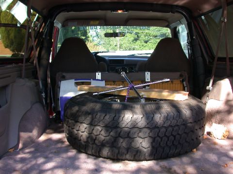 1996 Ford Bronco - Looking forward into the open cargo area of an SUV, where a full-size spare tire lies flat on the carpeted floor with a cross-style lug wre…