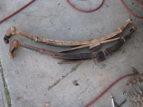 1978 Ford Bronco - Two aged leaf springs, one lighter and one darker, rest on a concrete floor with a red hose nearby.
