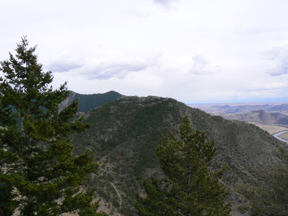 Mountainous terrain with scattered trees and a winding river under a cloudy sky