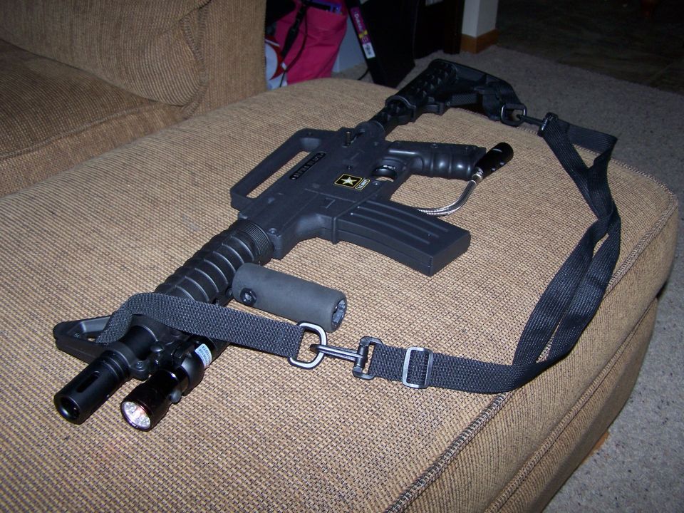 Black paintball gun with a sling and flashlight attachment resting on a couch
