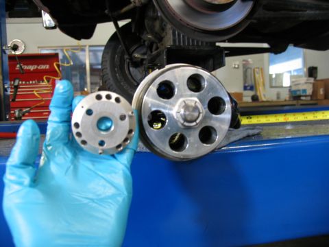 1984 Ford Bronco - A mechanic holds a pulley assembly next to a larger one, with a vehicle's undercarriage and suspension visible in the background.