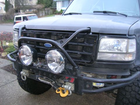 1996 Ford Bronco - Front view of a black Ford Bronco with an aftermarket bumper and spotlights, parked in a driveway with another vehicle behind it.