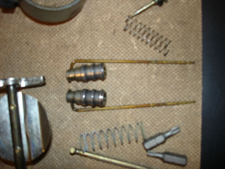 Carburetor components and springs laid out on a textured surface. A T-handle tool is also visible