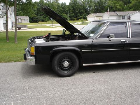 1987 Ford Crown Victoria LTD - The front end of a black sedan is visible with its hood open, revealing the engine compartment.