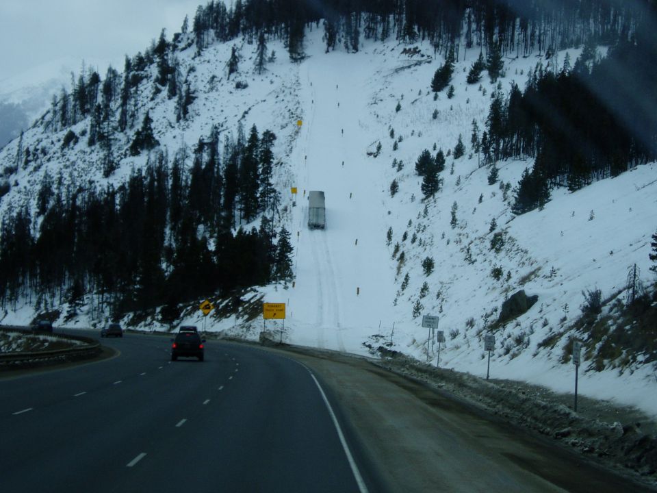 Dark sedan on highway with snow-covered mountain and truck on snowy road.