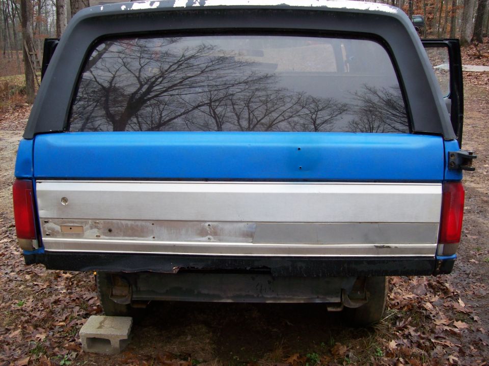 Rear view of a blue and silver Ford Bronco with a black camper shell, outdoors.