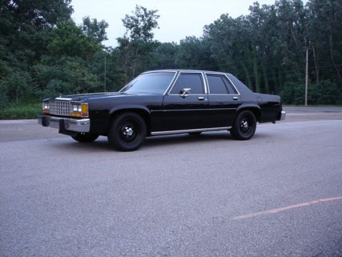 1987 Ford LTD Crown Victoria - A black four-door Ford LTD Crown Victoria sedan photographed from a front three-quarter angle, parked on an asphalt lot with green trees in…