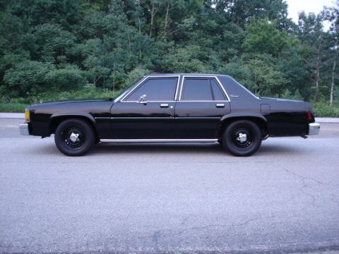1987 Ford LTD Crown Victoria - Full side profile of a black full-size four-door sedan parked on asphalt with dense green trees behind it. It rides on black steel wheels w…