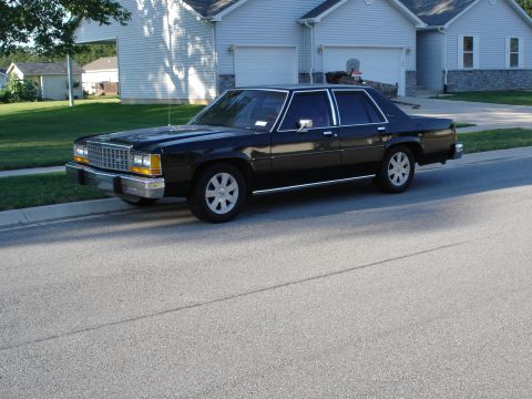 1987 Ford LTD Crown Victoria - Black boxy Ford LTD Crown Victoria four-door sedan parked in the street in front of a suburban two-car-garage house, shown from the front t…