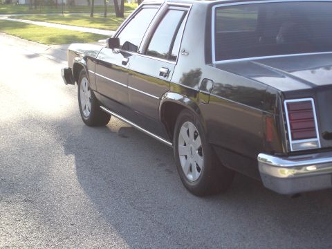 1987 Ford LTD Crown Victoria - Low rear three-quarter view of a black boxy Ford four-door sedan parked at a curb, showing white/silver five-spoke Ford police-style wheels…
