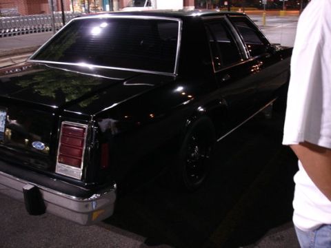1987 Ford LTD Crown Victoria - Night shot of a black four-door Ford full-size sedan from the rear three-quarter angle, showing vertical red tail lamps, chrome rear bumper…