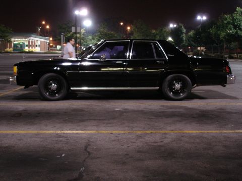 1987 Ford LTD Crown Victoria - Full side profile of a glossy black four-door full-size Ford sedan at night in a parking lot, sitting on black steel wheels with dog-dish c…