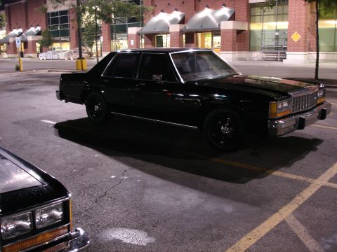 1987 Ford LTD Crown Victoria - Black boxy Ford four-door sedan parked at night in a store lot, shot from the driver's side front three-quarter with blacked-out steel whee…