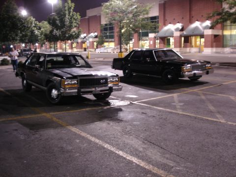 1987 Ford LTD Crown Victoria - Two black Ford full-size sedans parked nose-out at night in an empty retail parking lot, shot from the front three-quarter. The nearer car…