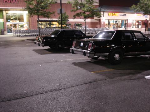 1987 Ford LTD Crown Victoria - Two black boxy Ford sedans parked nose-out in a strip-mall lot at night, shot from behind at a rear three-quarter angle; the near car wears…