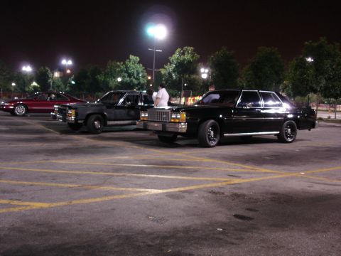 1987 Ford LTD Crown Victoria - Nighttime parking-lot meet with three cars lined up under light poles: a black boxy Ford four-door sedan on black wheels at right with its…