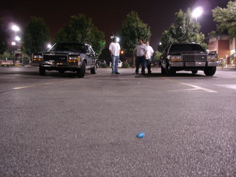 1987 Ford LTD Crown Victoria - Low ground-level night shot of two black full-size Ford sedans parked facing each other across an empty lot, both wearing chrome grilles, a…