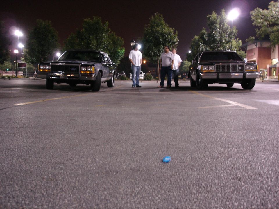 Two black full-size Ford sedans nose-out at night with three men standing between them
