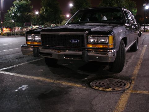 1987 Ford LTD Crown Victoria - Black boxy Ford four-door sedan photographed low and head-on at night in a parking lot, with a blue Ford oval centered in the dark eggcrate…