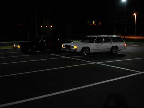 1987 Ford LTD Crown Victoria - Two station wagons are parked in a dark parking lot at night. The car on the left is black, and the car on the right is white.