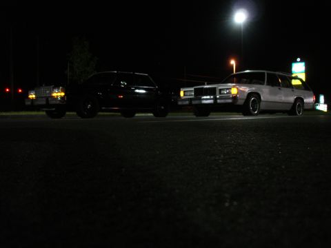 1987 Ford LTD Crown Victoria - Two station wagons are parked on asphalt at night. The car on the left is black and the car on the right is silver.