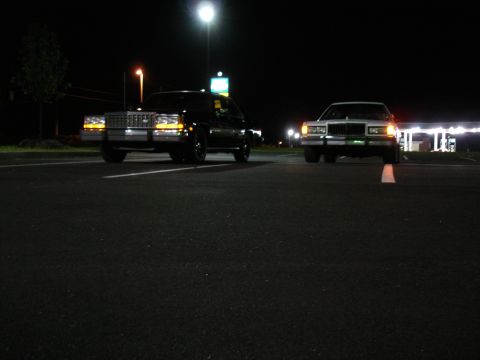 1987 Ford LTD Crown Victoria - Ground-level night shot across empty parking lot pavement of two full-size sedans facing the camera with headlights on. A black car with a…