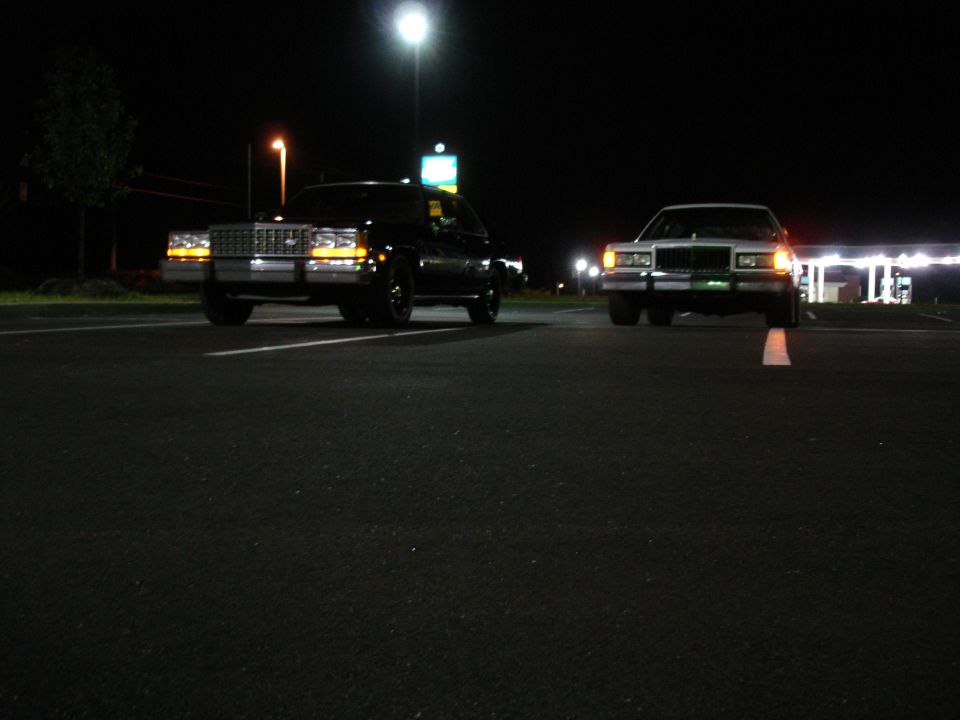 Two full-size sedans facing camera at night in a parking lot, headlights on, low angle