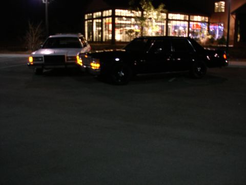 1987 Ford LTD Crown Victoria - Two sedans are parked in a lot at night in front of a building with illuminated windows. A white sedan is on the left, and a black sedan is…